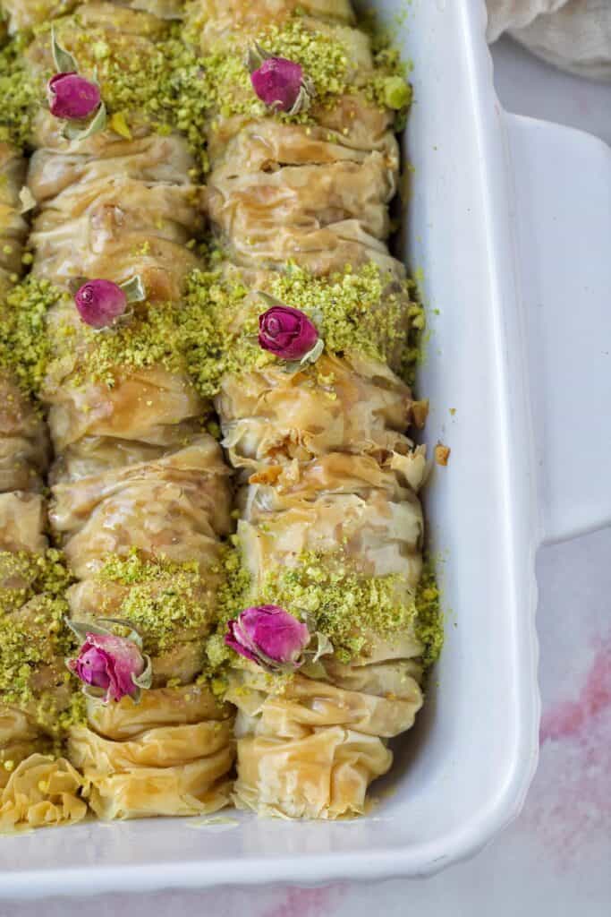 A beautiful dish of golden and flaky baklava rolls is sprinkled with pistachio and topped with little pink roses.