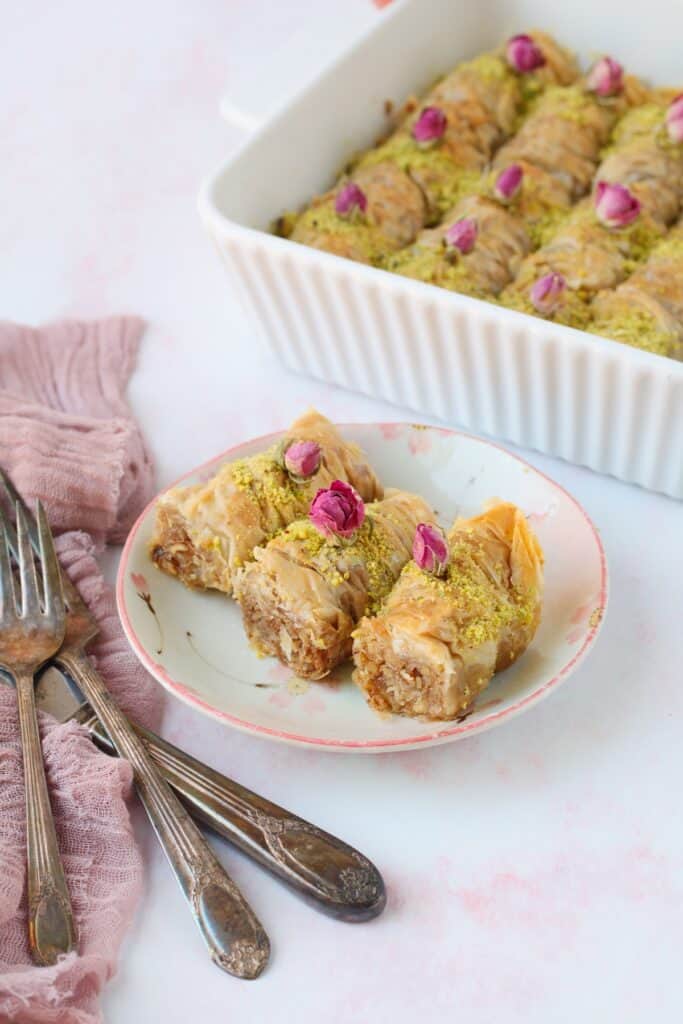 A small dish of crunchy rolled baklava filled with a chewy spiced walnut mixture, sprinkled with pistachio and decorated with dried pink roses.