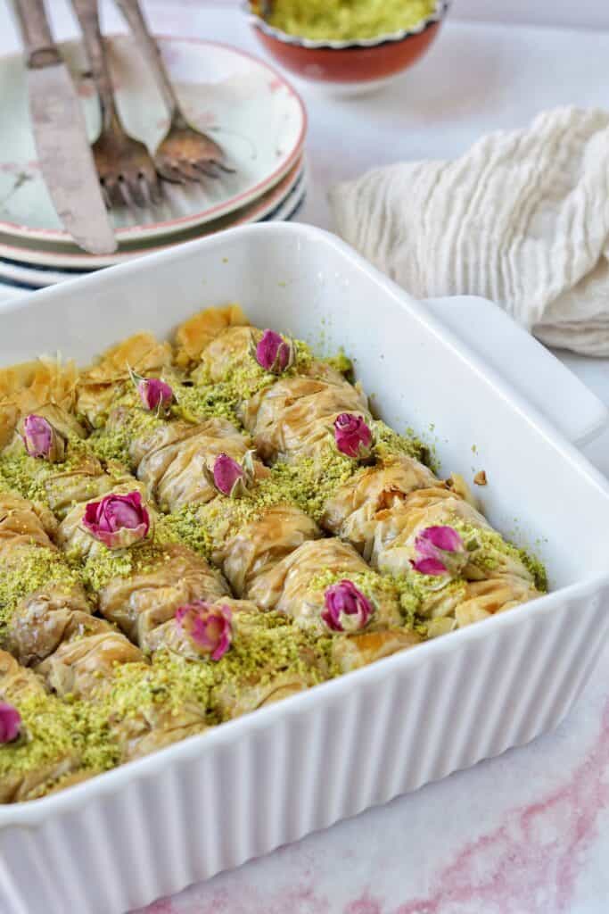 A beautiful dish of golden and flaky baklava rolls is sprinkled with pistachio and topped with little pink roses.