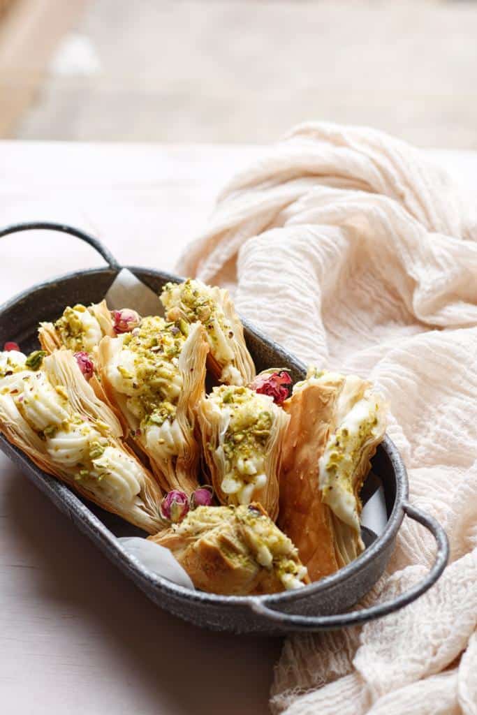 layers of crunchy phyllo dough, Shaabiyat are brushed with butter, filled with Ashta, garnished with crushed pistachios, drizzled with a syrup, and topped with rosebuds