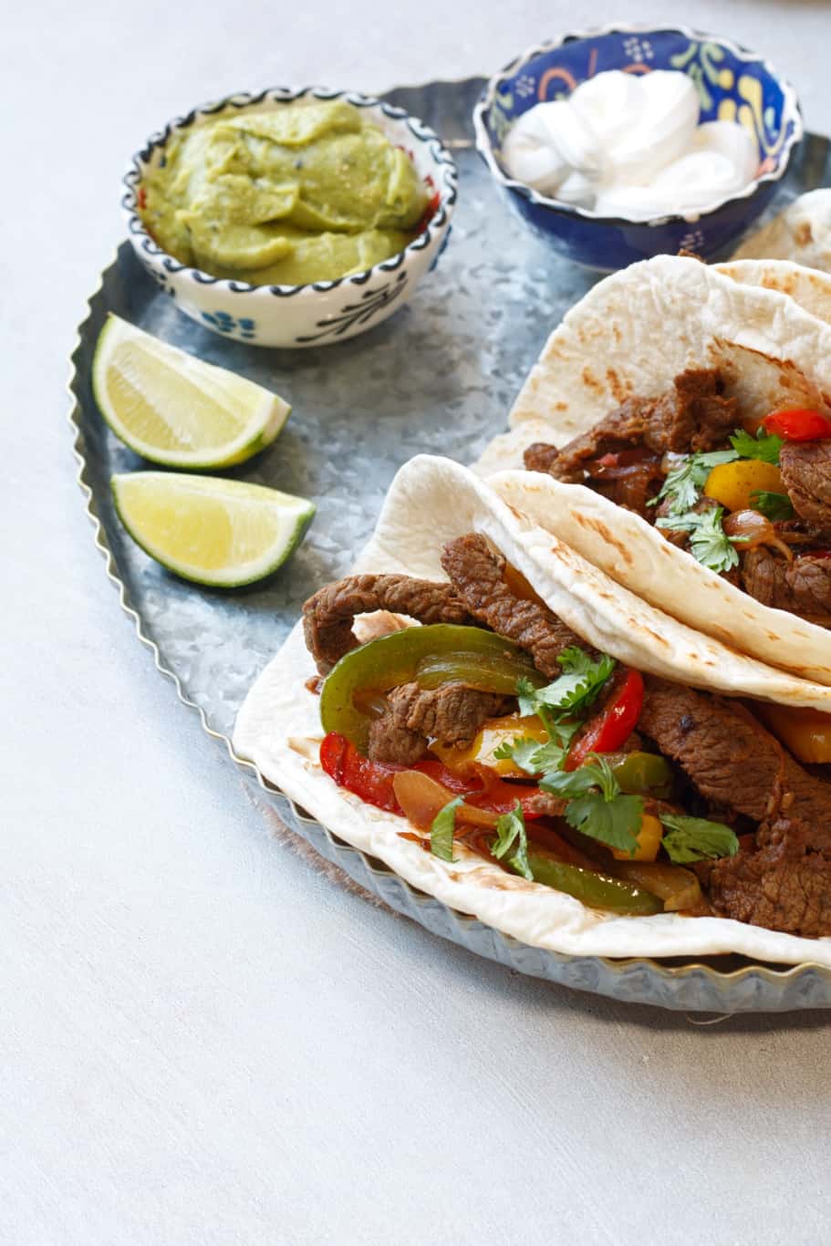 warm low-carb tortillas filled with keto beef fajitas which are made up of beef meat, onions, green, red, and yellow bell peppers and served with lime wedges, guacamole, and sour cream