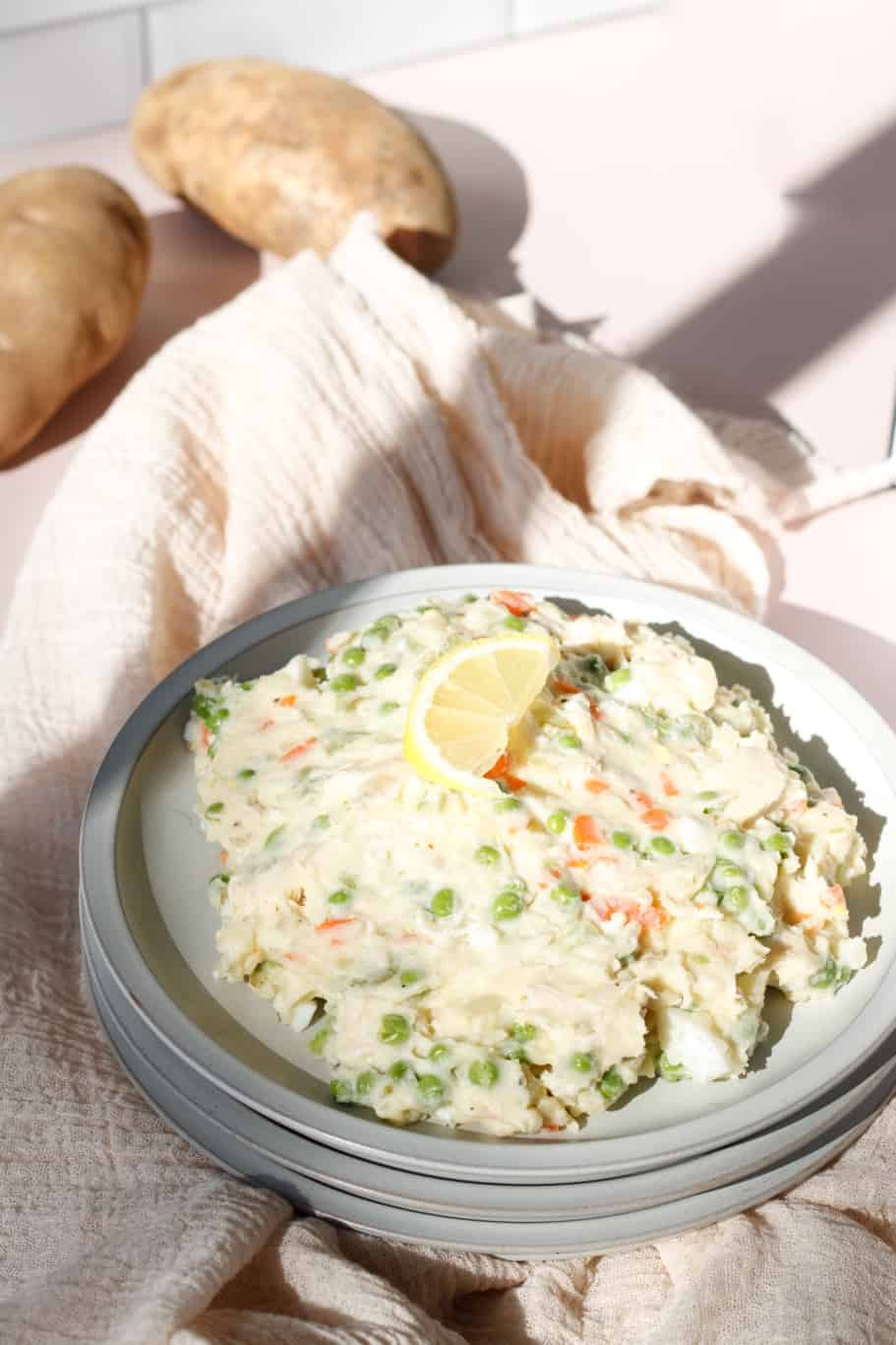 a plate of creamy tangy salad olivieh with a lemon wedge on the top. Alongside the dish, there are 2 medium russet potatoes.